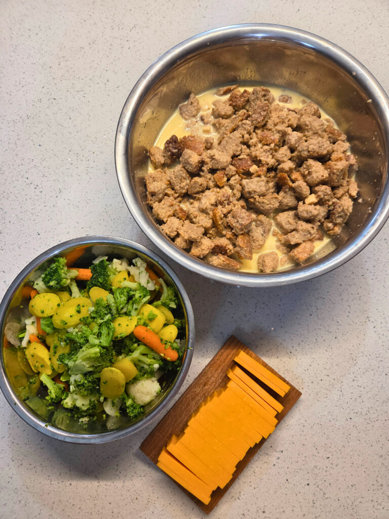 a bowl of mixed vegetables. a cutting board with cheese sliced, a bowl of bread soaking in milk and eggs