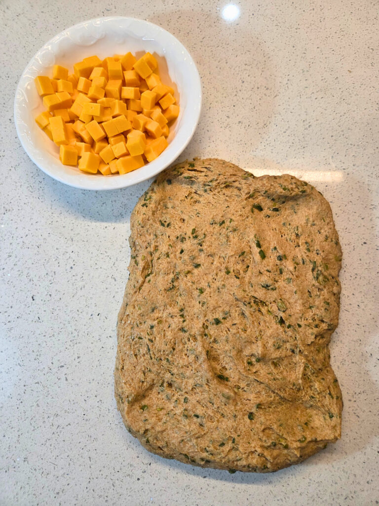 Flatten the Dough to add Cheese - Sourdough Cheddar and Chive Bread.jpg a flattened piece of dough next to a bowl of cheese cubes