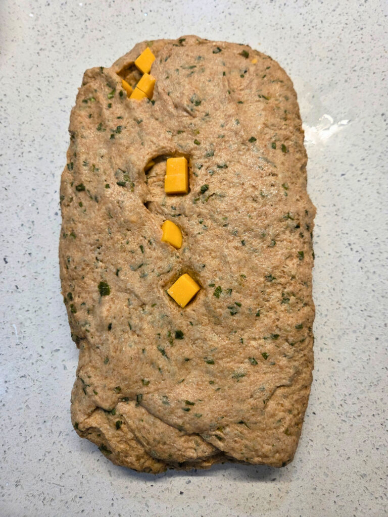 Fold Over the Edges - Sourdough Cheddar and Chive Bread.jpg a flattened piece of dough with cheese cubes sticking out from inside