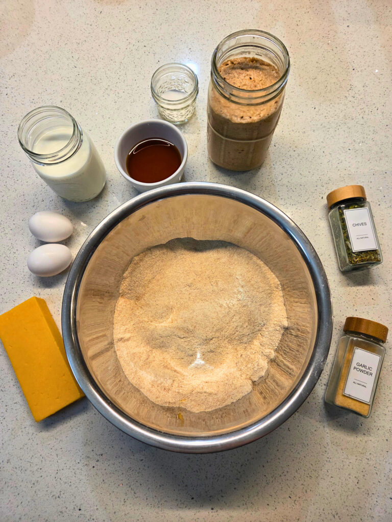 Ingredients for Sourdough Cheese and Chive Bread - Sourdough Cheddar and Chive Bread.jpg a bowl of flour, a cheese block, a jar of chives, a jar of garlic powder, 2 eggs, milk, honey, sourdough starter, and salt