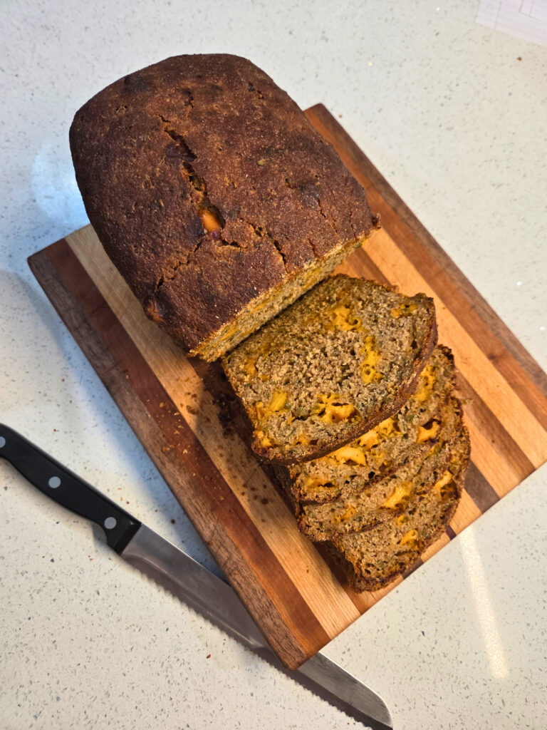 Slice the Loaf - Sourdough Cheddar and Chive Bread.jpg a partially sliced loaf of bread on a cutting board, next to a bread knife