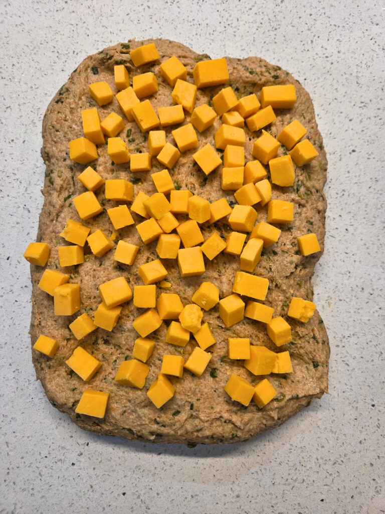 Sprinkle the dough with Cheese - Sourdough Cheddar and Chive Bread.jpg a flattened piece of dough with cheese cubes all over the top